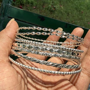 Op de afbeelding: Een verzameling zilveren armbanden in verschillende ontwerpen, waaronder gedraaid touw, kralen en ruitpatronen. De armbanden zijn op een hand gestapeld en tonen hun verschillende texturen en glans. De sieraden worden gepresenteerd tegen een groene achtergrond.
