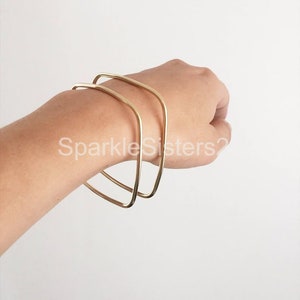 Puede incluir: Dos pulseras doradas cuadradas apiladas en la mu&ntilde;eca de una persona.