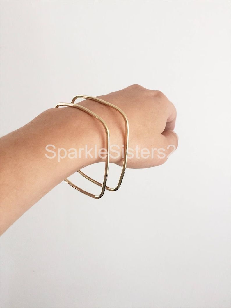 Puede incluir: Dos pulseras doradas cuadradas apiladas en la mu&ntilde;eca de una persona.