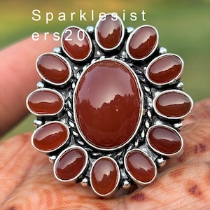 May include: A silver ring with a large, oval-shaped carnelian stone surrounded by smaller carnelian stones. The ring has a textured band.