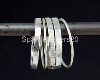 Op de afbeelding: Een verzameling zilveren ringen met gehamerde texturen. De ringen worden gepresenteerd op een metalen staaf tegen een donkere achtergrond. De ringen variëren in breedte en ontwerp en tonen een minimalistische esthetiek.