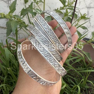 May include: Five silver bangles with intricate designs. The bangles are stacked on top of each other and have a variety of patterns, including floral, geometric, and rope-like designs.