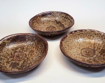 May include: Three brown ceramic bowls with a speckled glaze. The bowls are slightly oval shaped and have a raised design in the center.