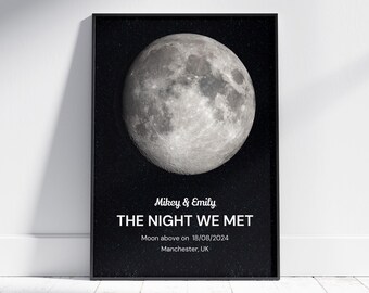 May include: Black framed print featuring a detailed grayscale image of the moon. The text "Mikey & Emily THE NIGHT WE MET" is displayed below the moon, along with the date 18/08/2024 and location Manchester, UK. The print is displayed on a white wall.