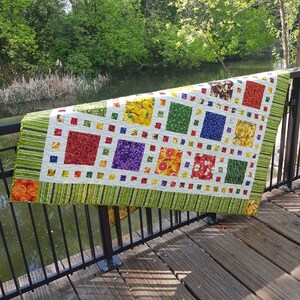 May include: A colorful quilt with a patchwork design featuring various fruits and vegetables. The quilt is made with a white background and green, red, yellow, and purple squares. The quilt is hanging on a black metal railing.