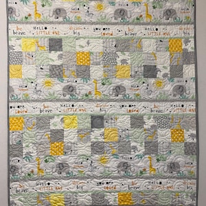May include: A gray and yellow baby quilt with a patchwork design featuring various animal illustrations, including giraffes, elephants, zebras, and suns. The quilt has the text "Hello Little One", "Dream Big", "You Are Loved", and "Be Brave" repeated throughout.