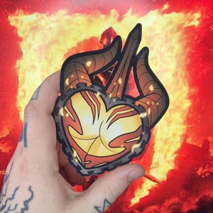 May include: A red and yellow heart-shaped sticker with a chain border and two horns. The sticker is on a hand in front of a fiery background.