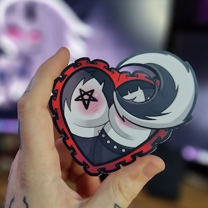 May include: A red heart-shaped sticker with a black and white illustration of two cartoon characters. The characters are a person with black hair and a pentagram on their cheek and a person with white hair. The sticker has a red lace border.