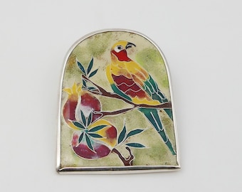 Op de afbeelding: Een zilveren broche met een kleurrijk emaille ontwerp van een vogel die op een tak zit met twee granaatappels. De vogel is geel, rood en groen, en de granaatappels zijn rood, geel en groen.