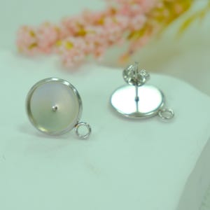 May include: Two silver earring components. One is a shallow, round dish with a small post in the centre and a loop on the side. The other is a flat, oval-shaped backing with a post and loop. Pink flowers are in the background.