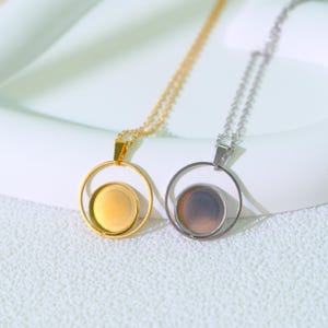 Könnte beinhalten: Zwei Halsketten mit runden Anhängern. Eine Kette hat eine goldfarbene Kette und einen Anhänger, die andere eine silberfarbene Kette und einen Anhänger. Die Anhänger haben einen kreisförmigen Rahmen und eine kleinere kreisförmige Basis.