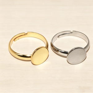 May include: Two adjustable rings, one gold-toned and one silver-toned, each featuring a circular, flat centerpiece. The rings are displayed on a light-colored surface, showcasing their simple, modern design. These rings are suitable for jewelry making.