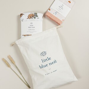 May include: A white muslin swaddle blanket with a brown bear and leaves print. The blanket is folded and placed in a white drawstring bag with the text "little blue nest" printed on it. The bag is sitting on a light beige surface.