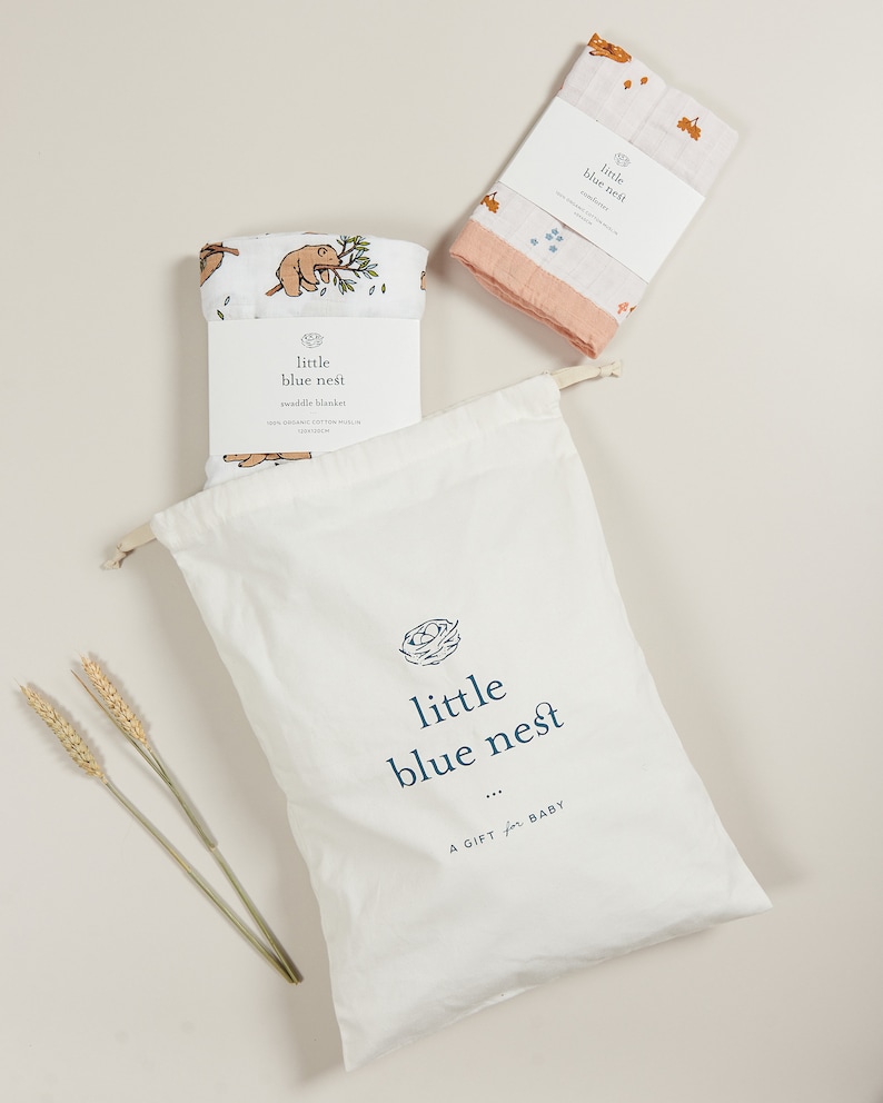May include: A white muslin swaddle blanket with a brown bear and leaves print. The blanket is folded and placed in a white drawstring bag with the text "little blue nest" printed on it. The bag is sitting on a light beige surface.
