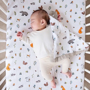 May include: A white cot sheet with a woodland animal print featuring foxes, squirrels, rabbits, and badgers. A baby is sleeping on the sheet, covered with a white muslin swaddle blanket with a similar print.