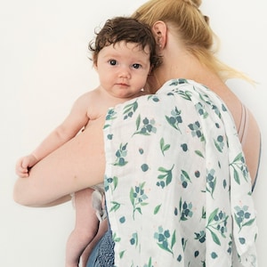 May include: A woman holding a baby wrapped in a white muslin swaddle with a green and blue floral pattern. The baby is looking at the camera.