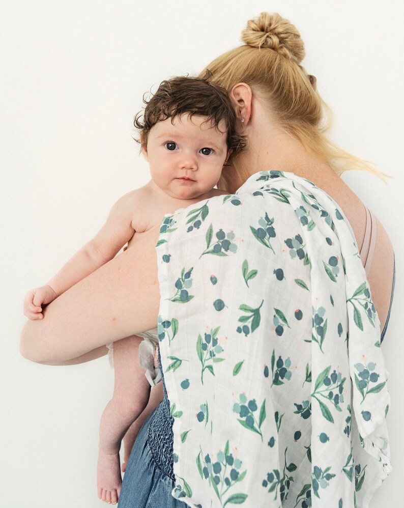 May include: A woman holding a baby wrapped in a white muslin swaddle with a green and blue floral pattern. The baby is looking at the camera.