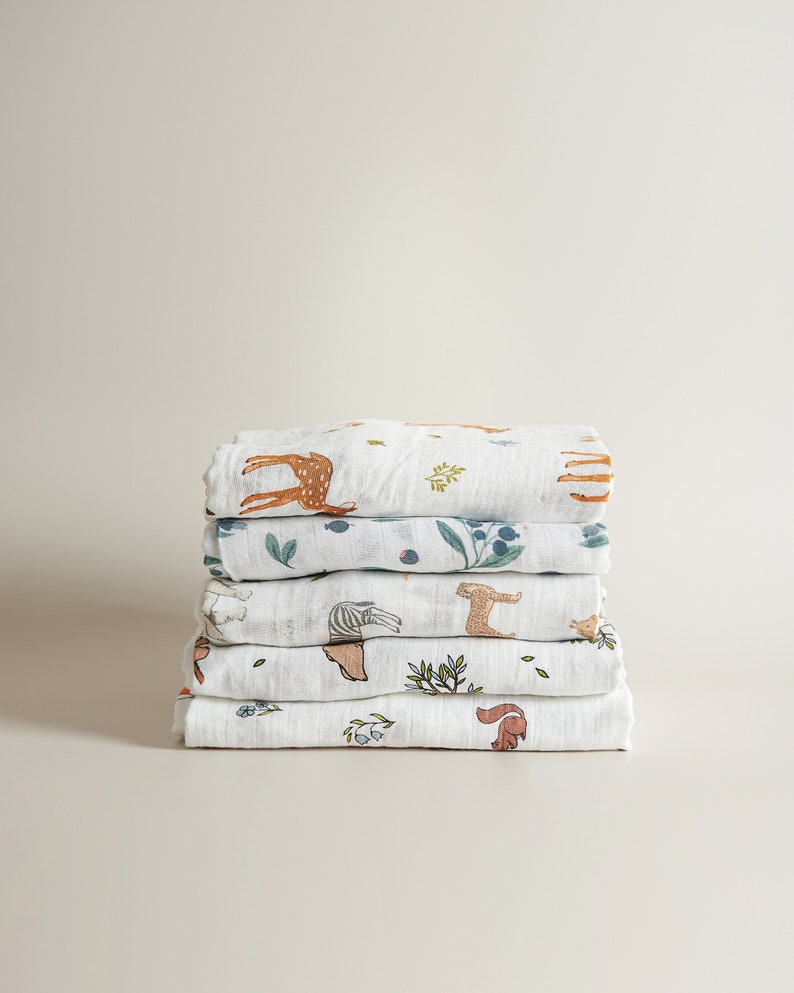 May include: A stack of six white muslin baby blankets with different animal and nature prints. The prints include a deer, zebra, squirrel, and leafy branches.