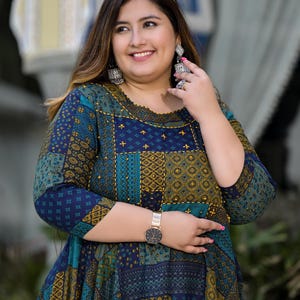 May include: A woman wearing a blue and green patterned dress with gold accents. She is wearing white sunglasses and silver earrings. She is also wearing a gold watch.
