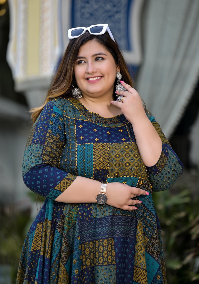 May include: A woman wearing a blue and green patterned dress with gold accents. She is wearing white sunglasses and silver earrings. She is also wearing a gold watch.