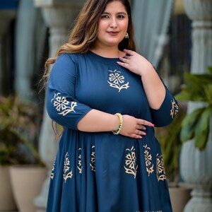 May include: A woman wearing a teal blue dress with a gold floral print. The dress has a round neckline and three-quarter sleeves. The dress is knee-length and has a flared skirt.