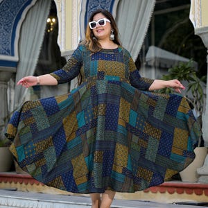 May include: A woman wearing white sunglasses and a blue and green patterned dress with gold accents. The dress has a flared skirt and is knee-length. She is standing in front of a building with arched doorways.