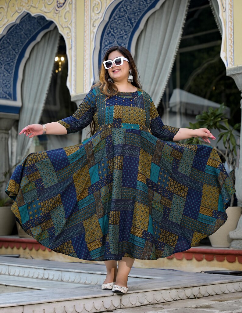 May include: A woman wearing white sunglasses and a blue and green patterned dress with gold accents. The dress has a flared skirt and is knee-length. She is standing in front of a building with arched doorways.