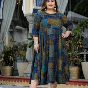 May include: A woman wearing a blue and green patterned dress with gold accents. The dress has a patchwork design and a flared skirt. She is wearing white sunglasses and white shoes.