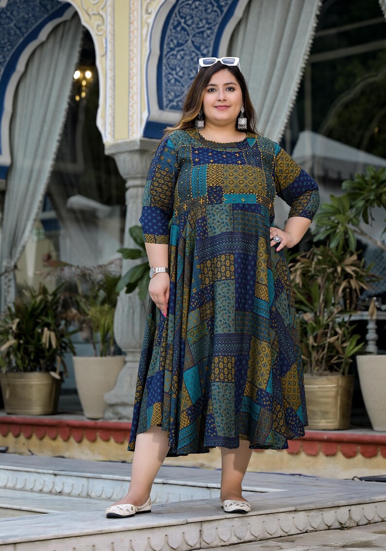 May include: A woman wearing a blue and green patterned dress with gold accents. The dress has a patchwork design and a flared skirt. She is wearing white sunglasses and white shoes.