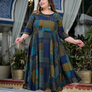 May include: A woman wearing a blue and green patterned maxi dress with a round neckline and three-quarter sleeves. The dress has a patchwork design with geometric shapes and floral patterns. The woman is standing on a stone step with a background of a building with arches.
