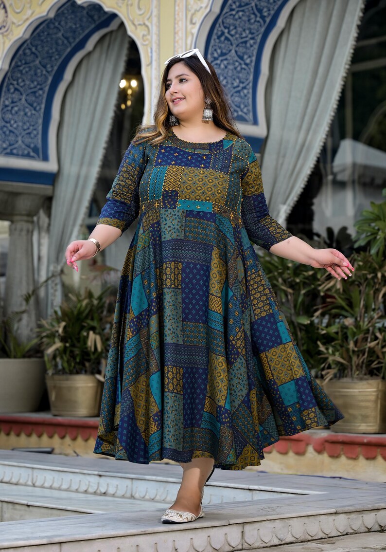 May include: A woman wearing a blue and green patterned maxi dress with a round neckline and three-quarter sleeves. The dress has a patchwork design with geometric shapes and floral patterns. The woman is standing on a stone step with a background of a building with arches.