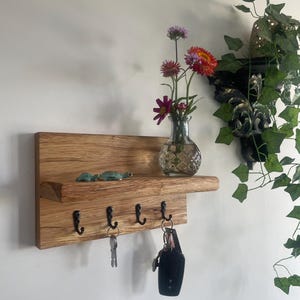 May include: A wooden key holder with four black hooks, a small shelf, and a decorative vase with flowers. Two small green turtle figurines sit on the shelf. Keys hang from the hooks.