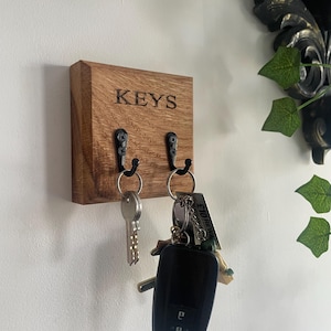 May include: Wooden key holder with the word "KEYS" engraved on the front. Two black metal hooks hold keys and keychains. The key holder is mounted on a white wall, with a decorative element and greenery in the background.