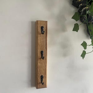 May include: A wooden key holder featuring three black metal hooks. The rectangular holder is crafted from light brown wood and is mounted on a white wall. The hooks are evenly spaced along the length of the wood.
