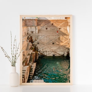 May include: A framed photograph of a large, ancient, stone structure with a pool of water at the bottom. The structure is made of many levels of steps and has a light brown color. There are people swimming in the pool of water.