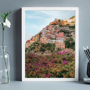 May include: A framed photograph of a colourful hillside village with a vibrant green foreground. The village is built into the hillside and features many buildings with terracotta roofs. The foreground is a lush green field with pink flowers.