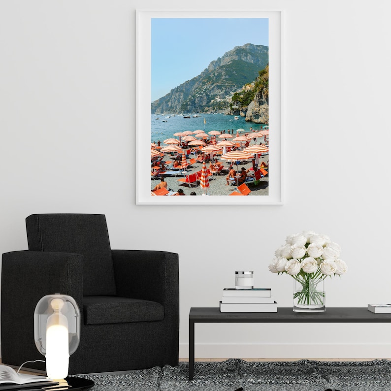 May include: A framed photograph of a beach scene with red and white striped umbrellas and people relaxing on the sand. The beach is located on a coastline with a mountain in the background.