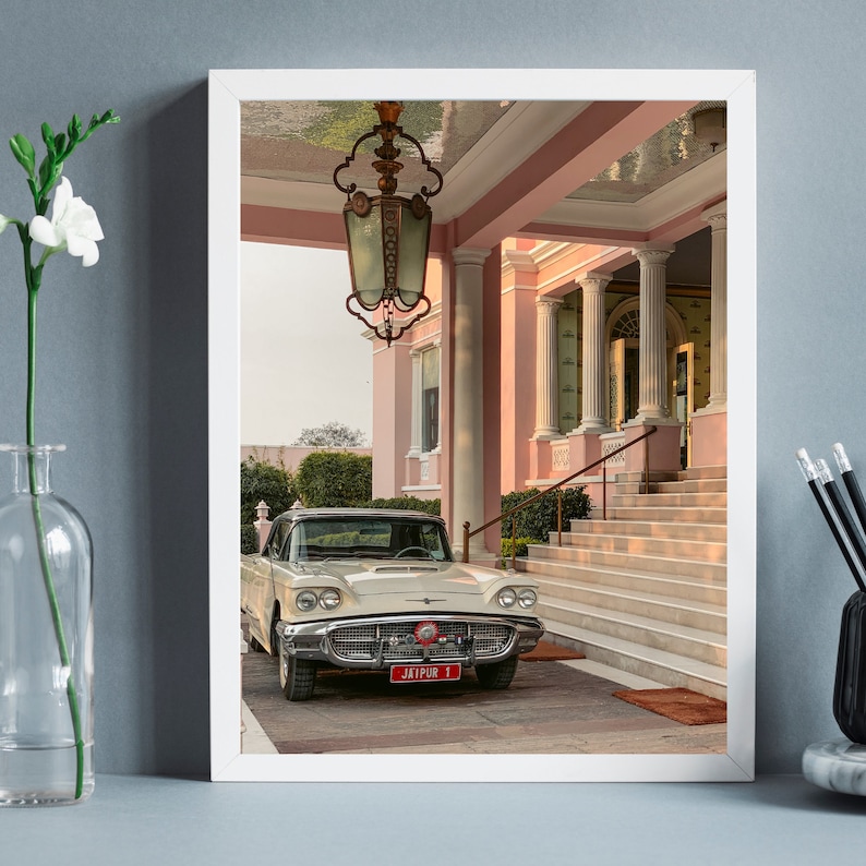 Op de afbeelding: Een witte vintage auto met een rood kenteken waarop "JAIPUR 1" staat, is geparkeerd voor een roze gebouw met een wit afdak. De auto is naar de camera gericht en het gebouw bevindt zich op de achtergrond.