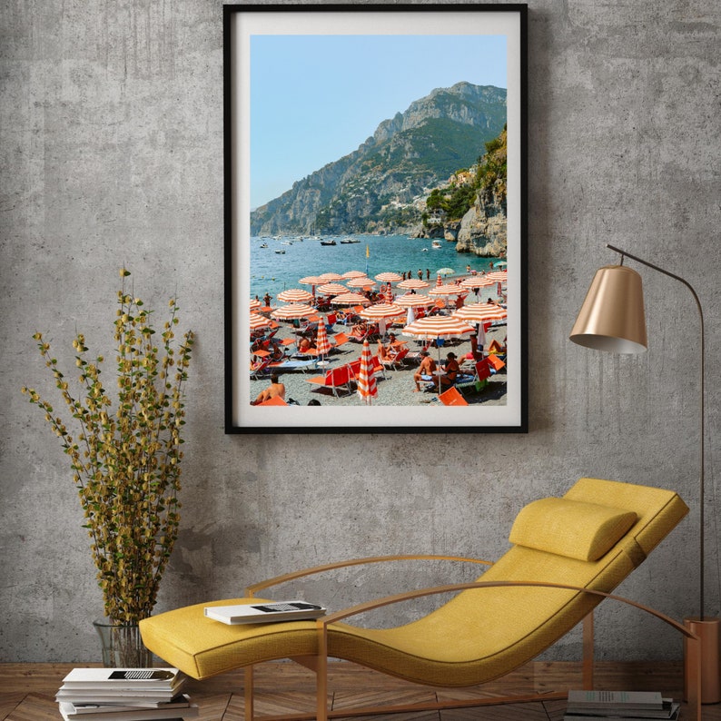 May include: A framed photograph of a beach scene with red and white striped umbrellas and people relaxing on the sand. The beach is located on a rocky coastline with a mountain in the background. The water is blue and the sky is clear.