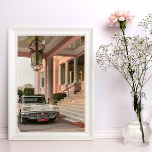 Op de afbeelding: Een vintage witte auto geparkeerd voor een roze gebouw met een grote, sierlijke kroonluchter die aan het plafond hangt. De auto heeft een rood kenteken met de tekst "JAIPUR 1".