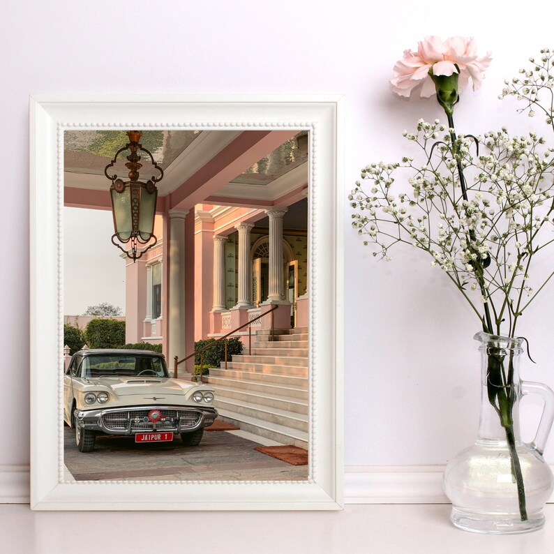 Op de afbeelding: Een vintage witte auto geparkeerd voor een roze gebouw met een grote, sierlijke kroonluchter die aan het plafond hangt. De auto heeft een rood kenteken met de tekst "JAIPUR 1".