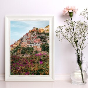 May include: A framed photograph of a colourful hillside town with a lush green foreground. The town is built on a steep slope and features many buildings with terracotta roofs. The foreground is filled with vibrant pink flowers.