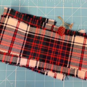 May include: A length of red, green, white, and navy blue tartan fabric with frayed edges. A small, red, berry-shaped embellishment is attached with twine. The fabric is on a blue cutting mat.