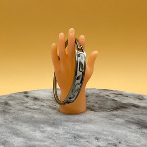 Peut inclure: Un bracelet jonc articulé argenté avec un motif incrusté blanc et irisé. Le bracelet est présenté sur un petit présentoir en forme de main orange sur fond jaune.