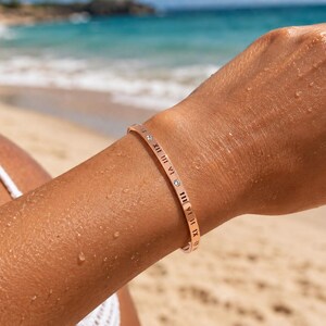 May include: A rose gold bangle bracelet featuring engraved black Roman numerals and small, clear, round-cut stones set at intervals along the outer surface of the thin, polished metal band, which encircles the wrist.