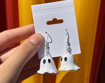 Op de afbeelding: Witte spookoorbellen met zwarte ogen en zilveren haken. De oorbellen worden gepresenteerd op een witte kaart tegen een achtergrond van oranje, geel en rood.