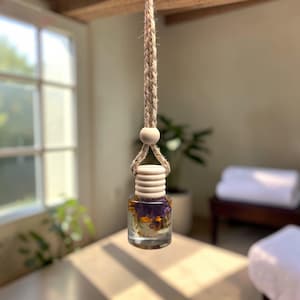 May include: A small, clear glass bottle filled with dried flowers and a light-colored wooden cap. The bottle is suspended by a natural fiber rope with a wooden bead. The background is a soft-focus interior with natural light.