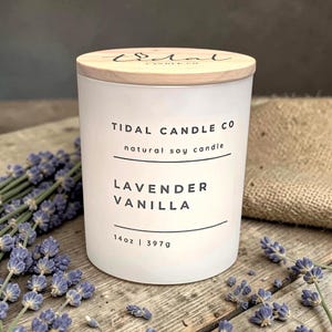 May include: A white glass candle with a wooden lid, labeled "Tidal Candle Co." and "Lavender Vanilla." The candle is 14 oz (397g). Lavender sprigs and a burlap sack are in the background, on a wooden surface.