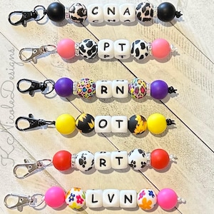 May include: Five colorful beaded keychains with different medical abbreviations. The keychains are made with black, pink, purple, yellow, and red beads with white letters spelling out CNA, PT, RN, OT, and RT. The keychains are on a light wood background.