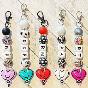 May include: Five colorful beaded keychains with a stethoscope charm. Each keychain features a different medical abbreviation:  'NP', 'RN', 'PA', 'RT', and 'RCP'.